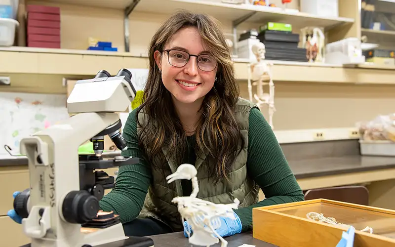 Student in a lab setting with a microscope and bird skeleton model, wearing gloves and seated at a workbench with anatomical models in the background.