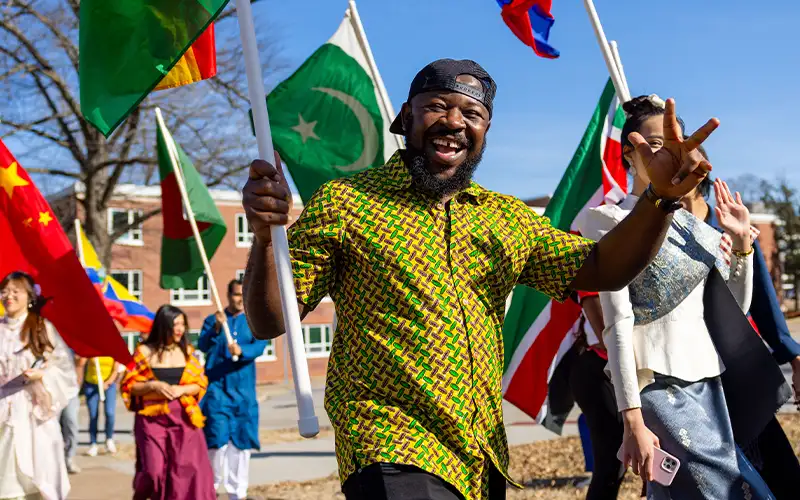 A smiling man in a patterned shirt holds a flag and flashes a peace sign during a multicultural Parade of Flags event, surrounded by participants carrying flags from various countries.